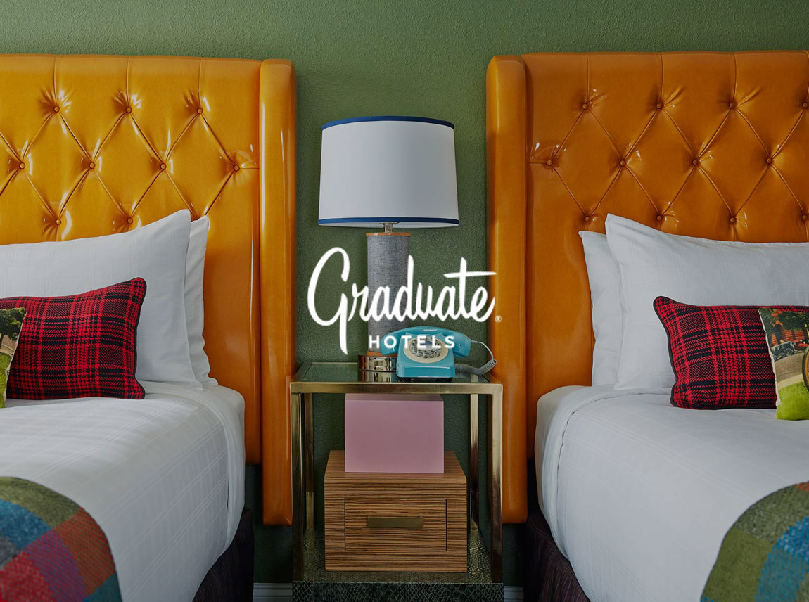A white Graduate Hotels logo on top of a hotel room with two queen beds and a side table sitting between them