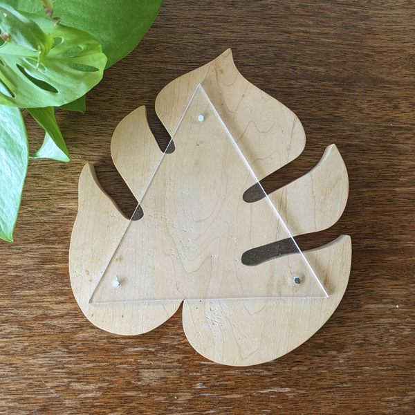A light wood cut piece in the shape of a monstera leaf with a triangle of plexiglass nailed to the top 